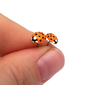 Pictured is a pair of orange ladybug earrings being held between a thumb and index finger for a close-up shot and size reference.