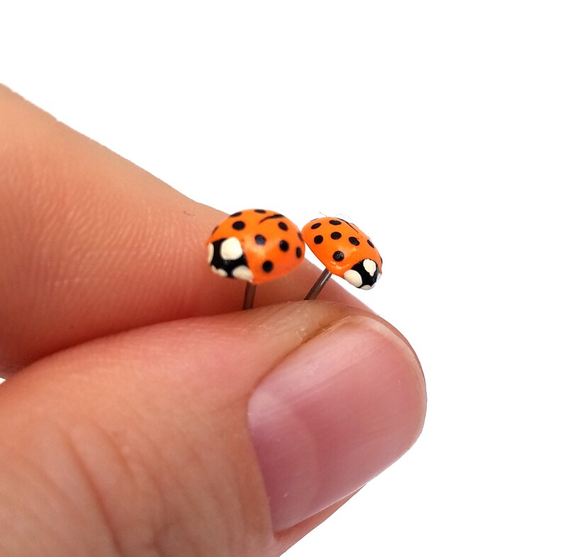 Pictured is a pair of orange ladybug earrings being held between a thumb and index finger for a close-up shot and size reference.