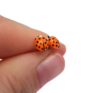 Pictured is a pair of orange ladybug earrings being held between a thumb and index finger to show another angle.