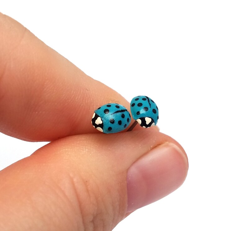 Pictured is a pair of light blue ladybug earrings being held between a thumb and index finger for a close-up shot and size reference.