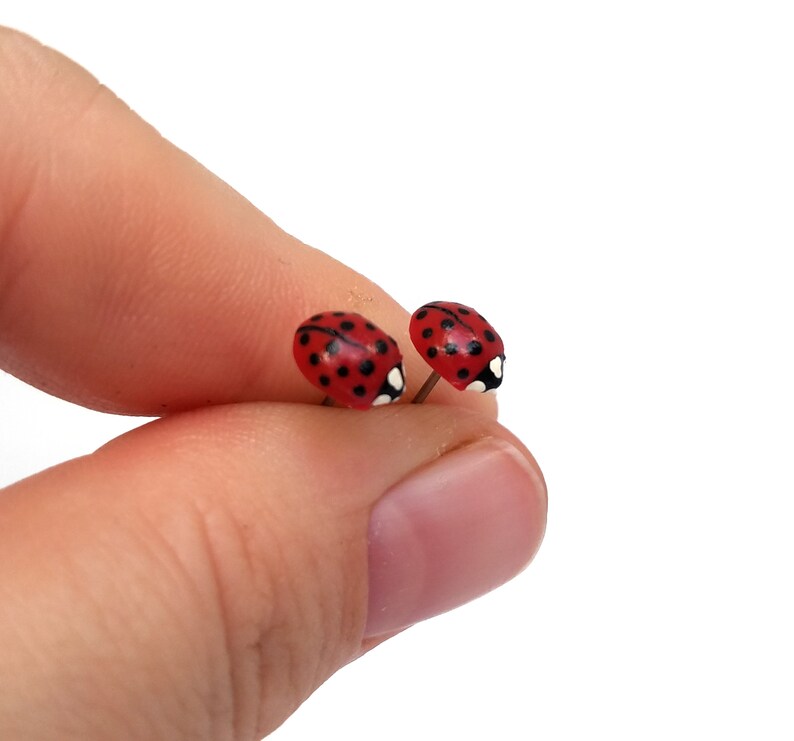 Pictured is a pair of realistic red ladybug earrings being held between a thumb and index finger for a close-up shot and size reference.