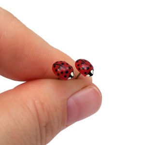Pictured is a pair of realistic red ladybug earrings being held between a thumb and index finger for a close-up shot and size reference.