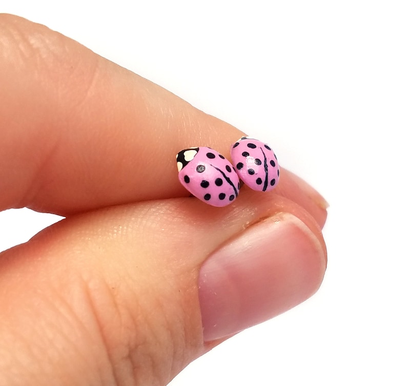 Pictured is a pair of pink ladybug earrings being held between a thumb and index finger for a close-up shot and size reference.
