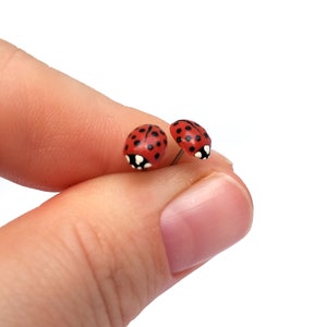 Pictured is a pair of realistic red ladybug earrings being held between a thumb and index finger for a close-up shot and size reference.
