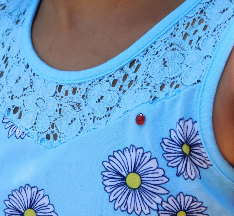 Pictured is a realistic, life-size ladybug pin pinned on a light blue summer dress with lace and daisies modeled by a girl with a medium brown skin tone.