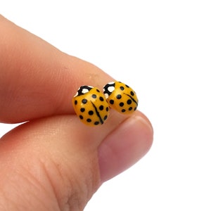 Pictured is a pair of yellow ladybug earrings being held between a thumb and index finger for a close-up shot and size reference.