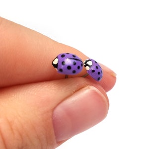 Pictured is a pair of purple ladybug earrings being held between a thumb and index finger for a close-up shot and size reference.