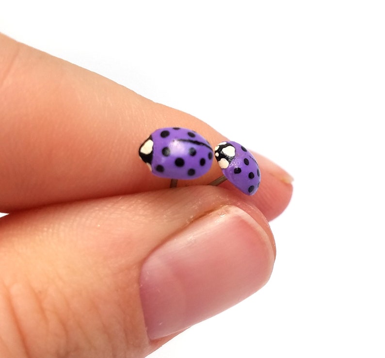 Pictured is a pair of purple ladybug earrings being held between a thumb and index finger for a close-up shot and size reference.