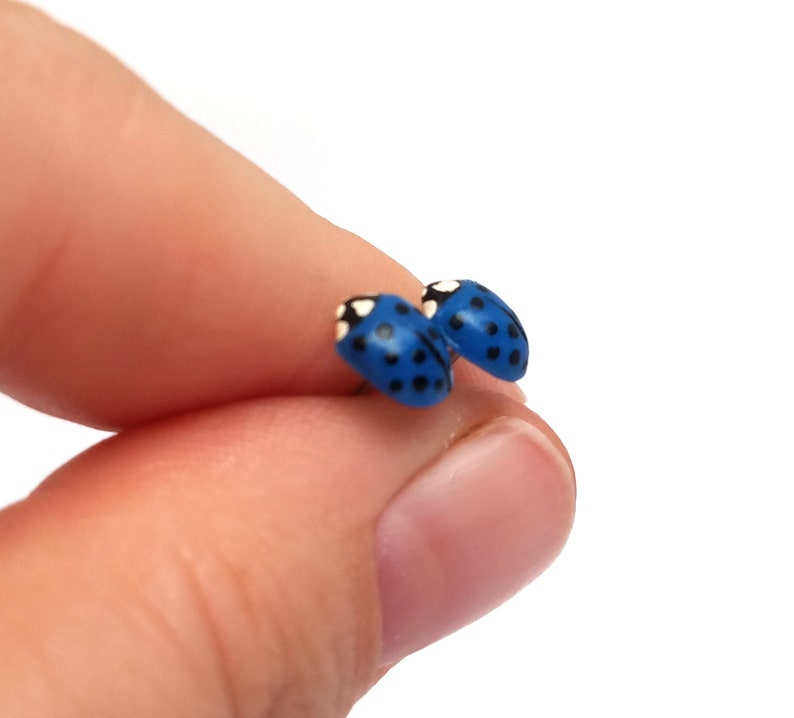 Pictured is a pair of dark blue ladybug earrings being held between a thumb and index finger for a close-up shot and size reference.