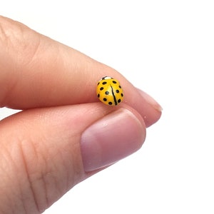 Pictured is a yellow ladybug pin being held between a thumb and index finger for a close-up shot and size reference.