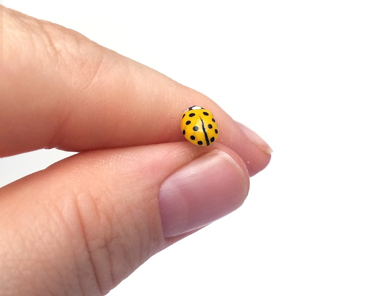 Pictured is a yellow ladybug pin being held between a thumb and index finger for a close-up shot and size reference.