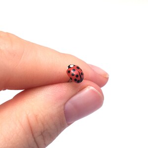 Pictured is a realistic ladybug pin being held between a thumb and index finger for a close-up shot and size reference.