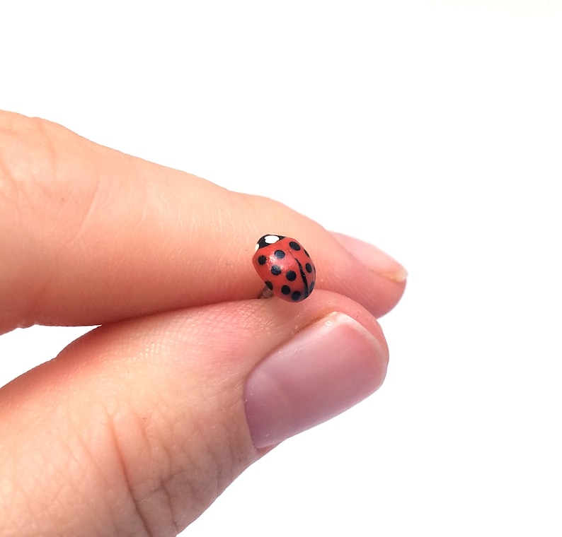 Pictured is a realistic ladybug pin being held between a thumb and index finger for a close-up shot and size reference.