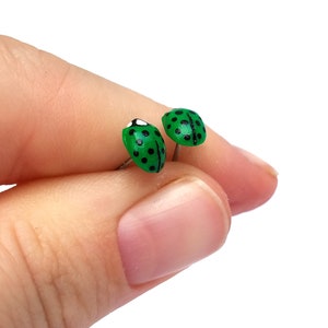 Pictured is a pair of green ladybug earrings being held between a thumb and index finger for a close-up shot and size reference.