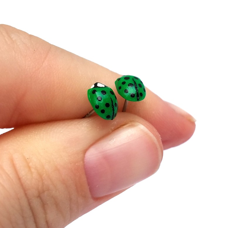 Pictured is a pair of green ladybug earrings being held between a thumb and index finger for a close-up shot and size reference.