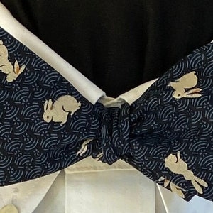 May include: A navy blue bow tie featuring a repeating pattern of cream-colored rabbits and a wave-like design. The bow tie is knotted and set against a white collared shirt and a black shirt.