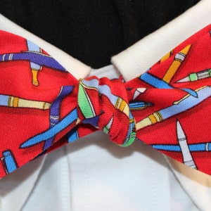 May include: A red bow tie featuring a repeating pattern of colorful pens in blue, yellow, and green. The bow tie is knotted and set against a white collared shirt.