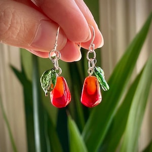 May include: Handmade earrings with a red and orange teardrop design, complemented by a green leaf. The earrings hang from silver-coloured hooks. The earrings are held up by a person's hand.