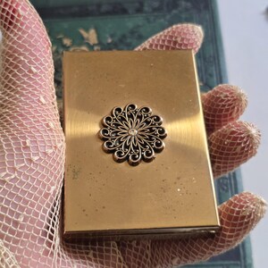 May include: A vintage gold-toned compact with a decorative floral design in the center. The compact has a rectangular shape and a hinged lid. The floral design is detailed with a central crystal. The compact is held by a hand wearing a lace glove.