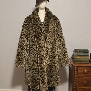 May include: A faux fur leopard print coat with a wide collar and long sleeves. The coat is displayed on a mannequin, paired with a brown cloche hat. The coat is knee-length and has a classic, vintage style.