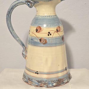 May include: A ceramic pitcher with a light blue handle and spout. The body is cream-colored with blue stripes and floral designs. The pitcher has a wide base and a decorative rim, perfect for serving beverages.