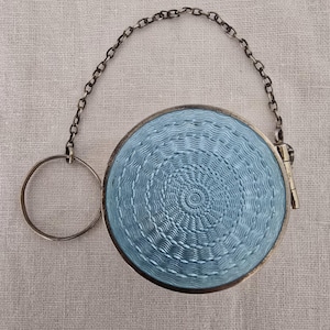 May include: A round, antique silver compact with a light blue, textured enamel design. The compact has a silver chain and a ring attached. The enamel pattern is a series of concentric circles. The compact is closed with a small clasp.