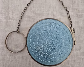 Könnte beinhalten: Ein rundes, hellblaues Puderdöschen mit einem texturierten, radialen Muster im Guilloché-Stil. Das Döschen ist an einer dünnen, silberfarbenen Metallkette befestigt, die in einen kreisförmigen Fingerring übergeht und als Armband dient.