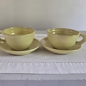 May include: Two matching ceramic cups and saucers in a pale, buttery yellow glaze. Each cup features a rounded bowl shape with a thick, curved handle attached to the side. The saucers are circular with a smooth, slightly raised rim design.