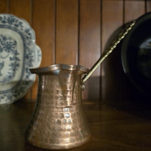 May include: A copper coffee pot with a long, decorative handle. The pot has a hammered texture and a flared base. The handle is gold-colored. The pot is sitting on a wooden surface.