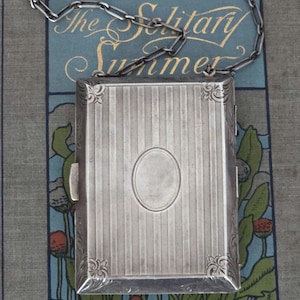May include: A rectangular silver-toned metal cigarette case featuring a vertical pinstripe pattern, an oval central cartouche, and decorative floral corner engravings. The case includes a silver-toned metal chain attached to the top edge.