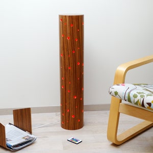 May include: A wooden floor lamp with a cylindrical shade featuring a wood grain pattern and red LED lights shining through small holes. The lamp is next to a chair with a floral patterned fabric and a wooden magazine rack.