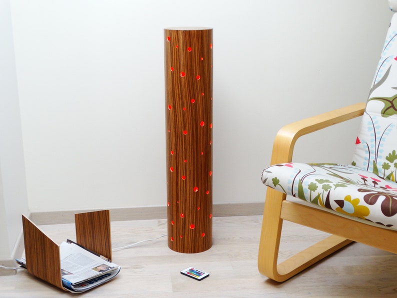 May include: A wooden floor lamp with a cylindrical shade featuring a wood grain pattern and red LED lights shining through small holes. The lamp is next to a chair with a floral patterned fabric and a wooden magazine rack.