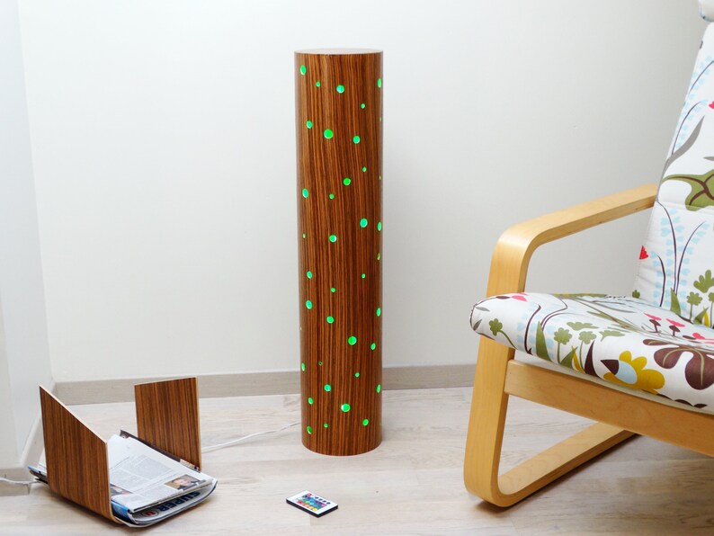 May include: A wooden floor lamp with a cylindrical base and green LED lights shining through holes in the wood. The lamp is next to a wooden chair with a floral patterned cushion.