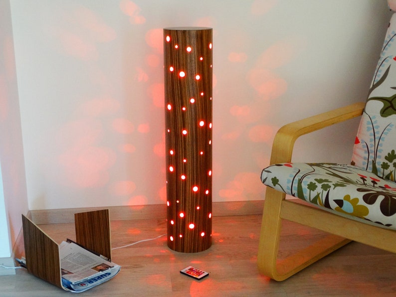 May include: A wooden floor lamp with a cylindrical base and a pattern of holes. The lamp is lit with red light, casting a warm glow on the surrounding area. A remote control is on the floor next to the lamp.
