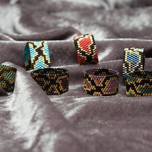 Può includere: Una collezione di otto anelli di perline con vari motivi geometrici e astratti in nero, oro, blu, verde e rosso.