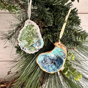 May include: Two handmade Christmas ornaments shaped like oyster shells. One is painted with green and blue swirls and the other is painted with blue and white swirls. Both have gold accents.