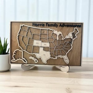 May include: A wooden map of the United States with state outlines and the text "Norris Family Adventures." The map is displayed on a wooden stand. States of Colorado, Kansas, Texas, and Florida are labeled.