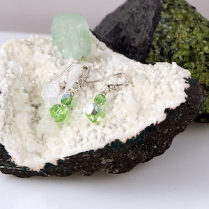 May include: A pair of silver earrings with green and white crystal beads. The earrings are displayed on a white rock with a green rock in the background.