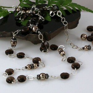 May include: A long necklace featuring a combination of dark brown, oval-shaped beads and smaller, round, pearl-like beads. The beads are linked together with silver-colored chain. The necklace is displayed on a dark, textured surface.
