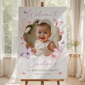 May include: A white sign on an easel, featuring a baby girl's photo and butterfly illustrations. The sign's text reads "Welcome to Caroline's 1st Birthday Party, February 27, 20XX" in a soft purple script. Butterflies in pink and purple.