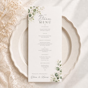 Puede incluir: Una tarjeta de menú de cena vertical con las palabras "The Dinner Menu" en una elegante escritura, acentuada con hojas de eucalipto en acuarela. El menú enumera entrantes, sopa, platos principales, postre y bebidas. Los nombres "Olivia & Marcus" están en la parte inferior.