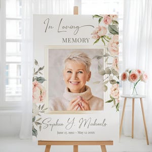 May include: A memorial sign on an easel features a photo of a woman, framed with gold lines. The sign reads "In Loving Memory" and includes the name "Sophia Y. Michaels" and dates. Floral accents in pink and green decorate the sign.