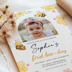 May include: A personalized invitation with a photo of a baby girl, adorned with a pink bow, celebrating her first birthday. The design features cartoon bees, honeycomb patterns, and the text "Sophia's first bee-day."