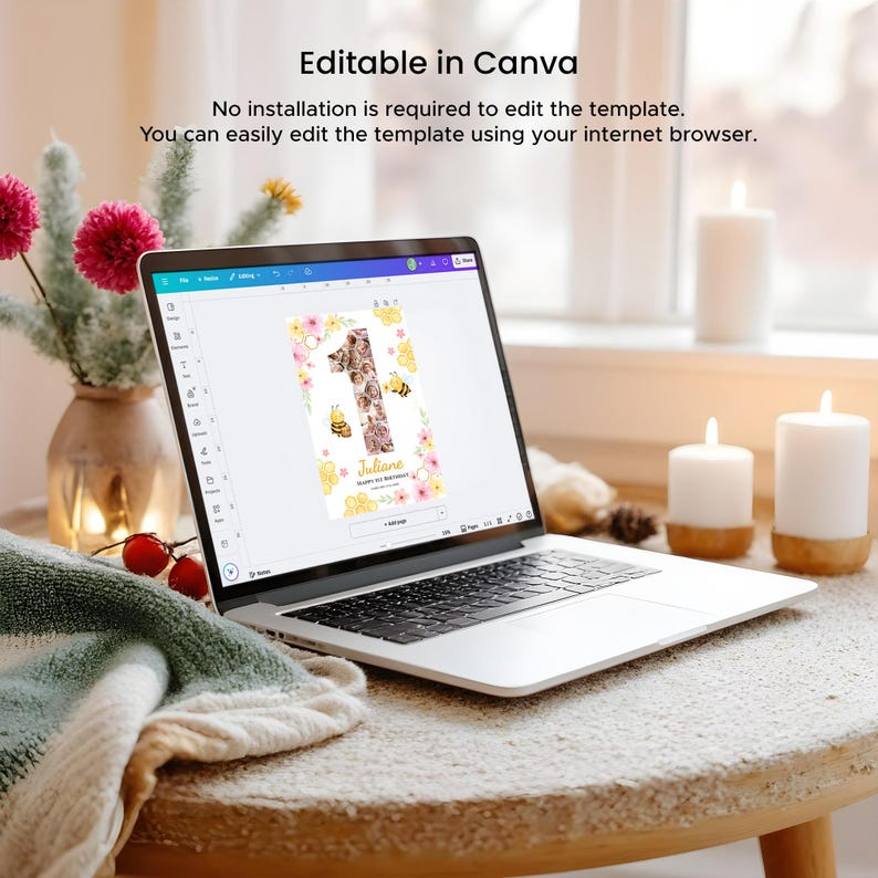 May include: A laptop displays a birthday invitation template with the number one, bees, and floral accents. The text "Editable in Canva" and instructions are visible. The laptop sits on a round wooden table with a cozy blanket and candles.