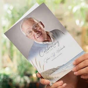 May include: A memorial card held by a person, featuring a portrait of an elderly man wearing glasses and a light blue sweater. The card reads "Celebrating the Life of John Y. Michaels" with a beach scene illustration.