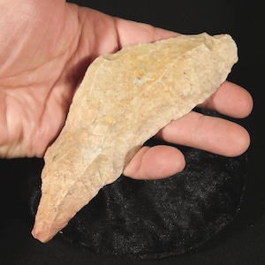 May include: A tan-colored, elongated stone tool with a pointed end and chipped, textured edges. The object features a rough, uneven surface with variations in light brown and beige tones, showcasing a sharp, tapered shape typical of a flint blade.