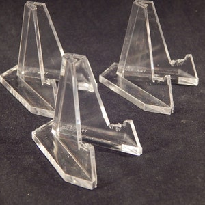 May include: Three clear acrylic display stands. Each stand has a triangular support and a rectangular base. The stands are designed to hold items upright for display purposes. The stands are on a dark background.