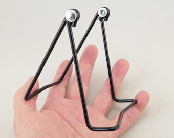 May include: Black metal display stand with a triangular shape. The stand has two metal hinges with silver accents.