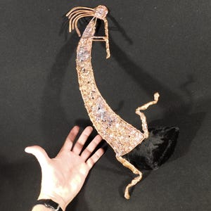 May include: A copper-colored metal sculpture of a flute player with a curved, textured body and a black base. The figure features a slender, arched form with wire-like details representing hair and limbs, finished with a metallic, reflective surface.
