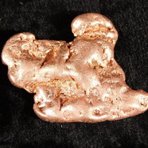 Könnte beinhalten: Ein roher, metallischer Kupferklumpen mit einer natürlichen, unregelmäßigen Form. Die Oberfläche zeigt einen rotbraunen Farbton mit einer Mischung aus glatten, abgerundeten Abschnitten und texturierten, porösen Bereichen, die das Licht reflektieren.